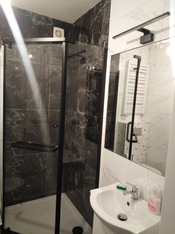 Nowoczesna łazienka po remoncie z kabiną prysznicową z czarnymi ramami i marmurkowymi płytkami w odcieniach szarości i bieli, widok na umywalkę i lustro z oświetleniem LED.