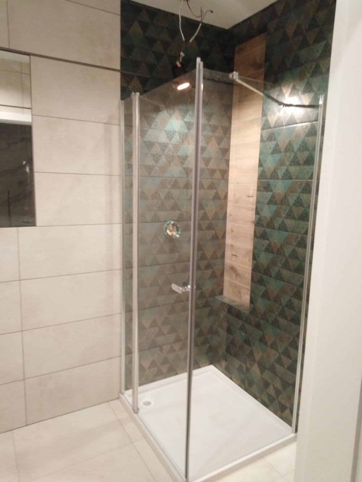 Kabina prysznicowa z brodzikiem, zamontowana w wykończonej łazience z geometrycznymi płytkami w odcieniach brązu i zieleni oraz jasnymi płytkami na sąsiedniej ścianie.