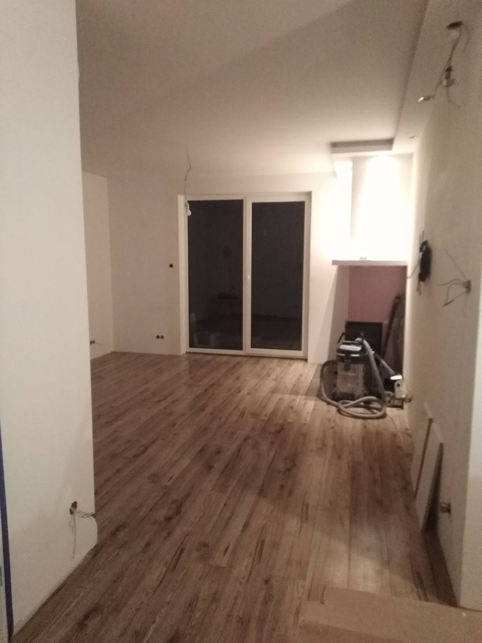 Wnętrze pokoju po remoncie z drewnianą podłogą, widoczny odkurzacz przemysłowy i niedokończone punkty elektryczne na ścianach.