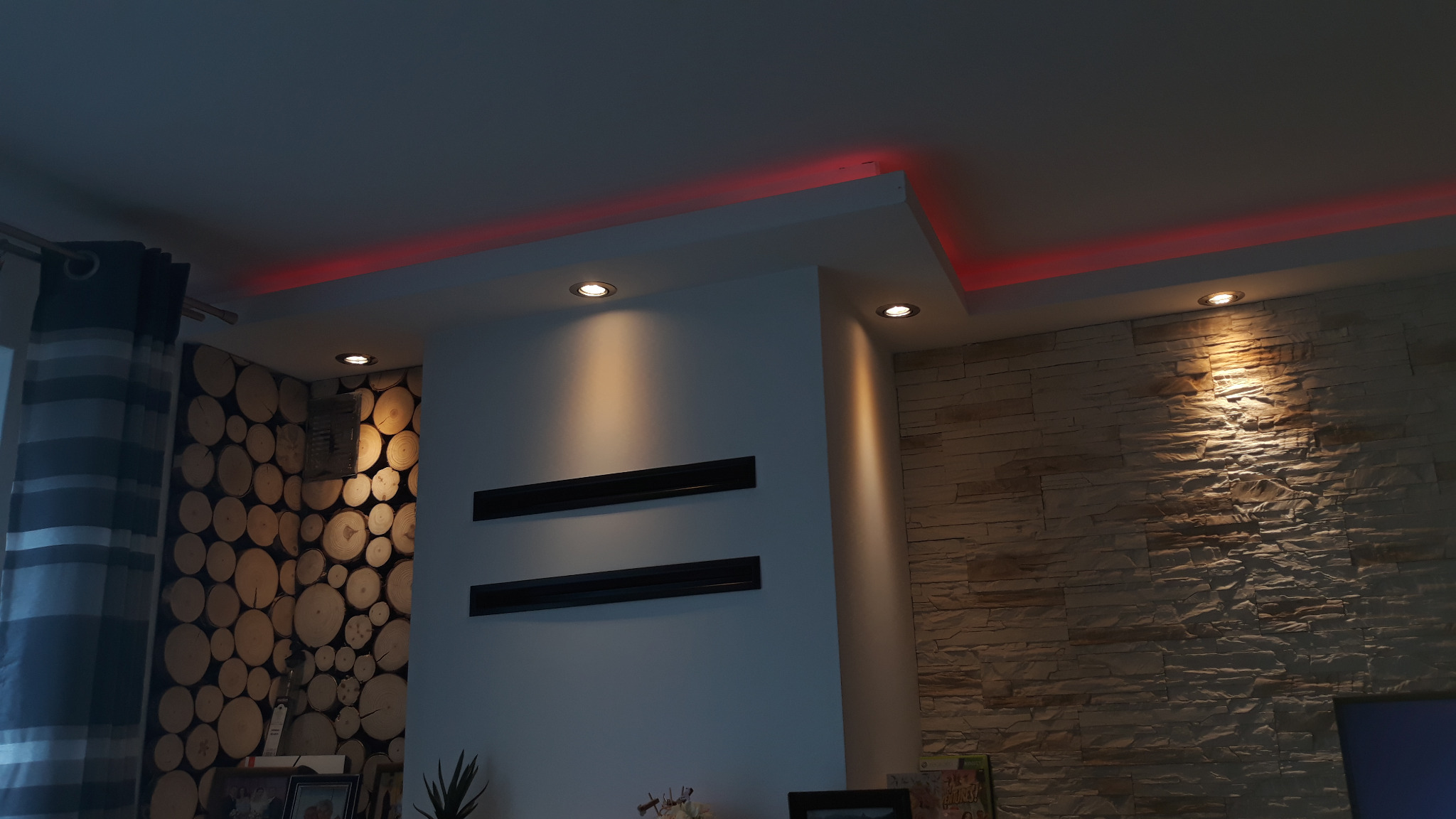 Nowoczesny salon z podwieszanym sufitem, oświetleniem LED w kolorze czerwonym i punktowymi lampami, dekoracyjną ścianą z drewnianych plastrów i kamienną okładziną.