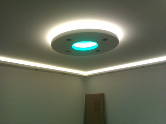 Nowoczesny sufit z oświetleniem LED: okrągła lampa wpuszczana z turkusowym światłem w centrum i listwy LED wzdłuż krawędzi sufitu, neutralne ściany, kartonowe pudło w rogu pokoju.