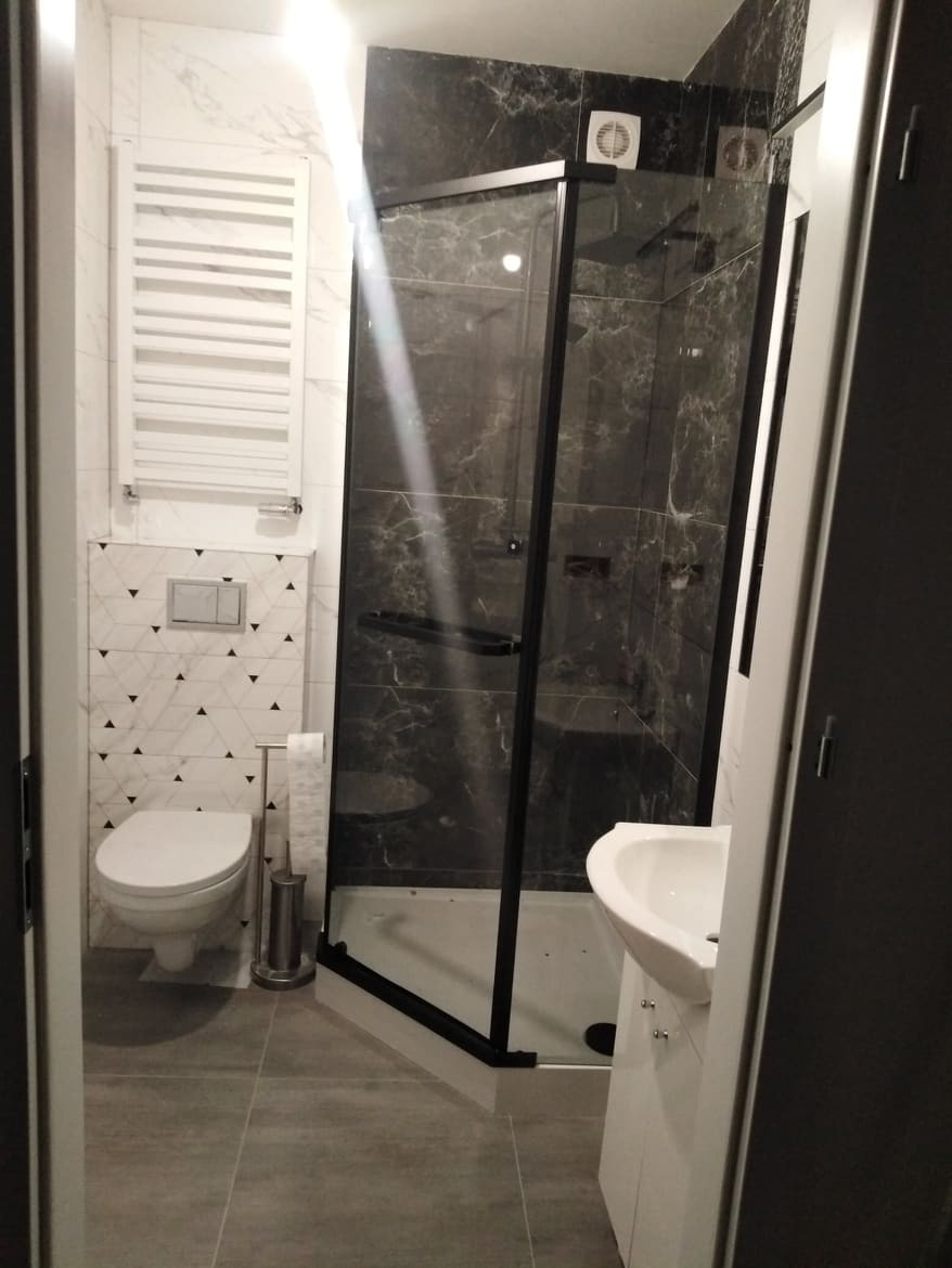 Nowoczesna łazienka po remoncie z kabiną prysznicową z czarnymi ramami, wiszącą toaletą i grzejnikiem drabinkowym na ścianie z geometrycznym wzorem.