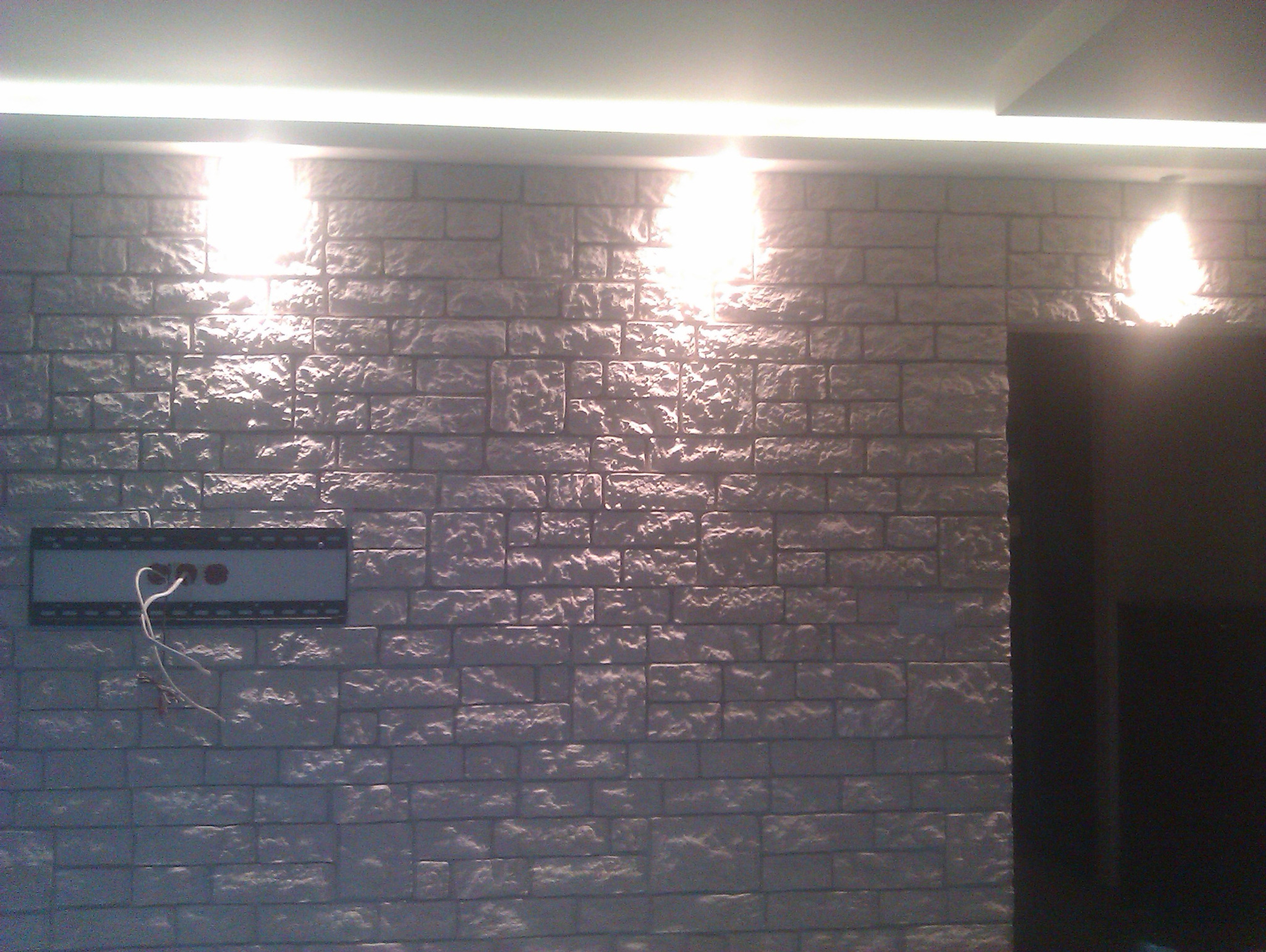 Ściana wykończona dekoracyjną cegłą gipsową w odcieniach szarości, oświetlona punktowo, z widoczną puszką elektryczną i wystającymi przewodami, obok ciemnego otworu drzwiowego.