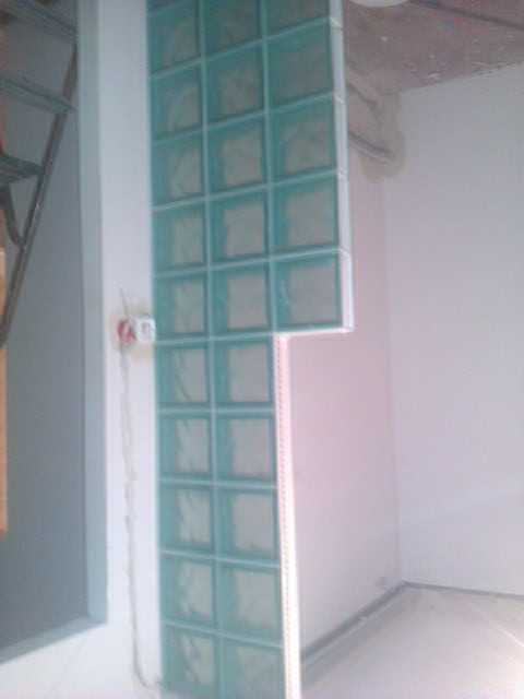 Ścianka działowa z luksferów w odcieniach błękitu, w trakcie montażu w narożniku pomieszczenia, widoczne niedokończone wykończenie sufitu i wystające przewody elektryczne.