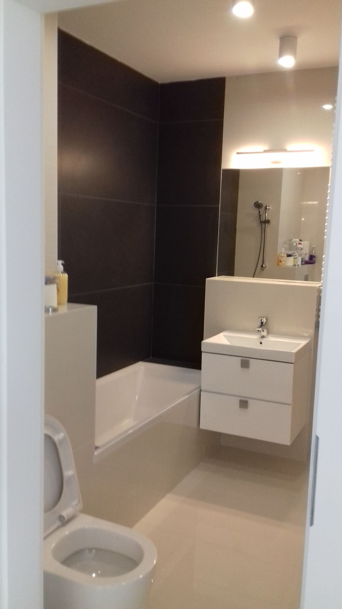 Nowoczesna łazienka z czarnymi płytkami na ścianie, białą wanną, wiszącą szafką z umywalką i toaletą z otwartą klapą.