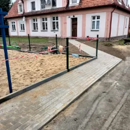 Ścieżka z kostki brukowej w trakcie budowy, obok piaskownica z elementami placu zabaw, w tle budynek z różową elewacją i czerwoną dachówką, teren ogrodzony taśmą ostrzegawczą.