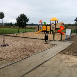 Nowo wybudowany plac zabaw z kolorowymi elementami zabawowymi, ogrodzony metalową siatką, otoczony żwirową nawierzchnią i młodym drzewkiem z kulistą koroną, przy częściowo utwardzonej drodze