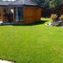 Widok na zadbany trawnik z dwoma drewnianymi altanami ogrodowymi; jedna z przeszkleniami i kwiatami w donicach, druga z kamienną podstawą i schodkami, również z kwiatami.