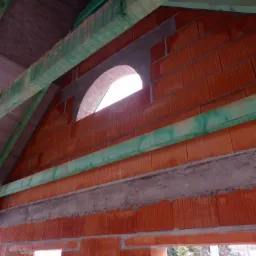 Wnętrze budynku w trakcie budowy; widoczna ściana z czerwonej cegły z otworem okiennym w kształcie łuku, zielone elementy konstrukcji dachu oraz betonowy nadproże.