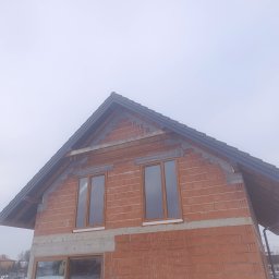 KRZYSZTOF HABRYŁO HAB-KRIS - Surowy stan domu z czerwonej cegły z zamontowanymi oknami i szarym dachem, widoczne elementy konstrukcyjne, pochmurne niebo w tle. Budowa domu w trakcie realizacji.