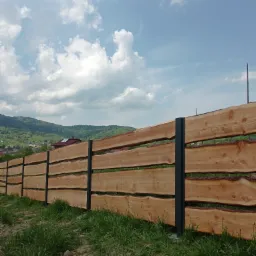 Drewniane ogrodzenie z poziomych desek o nieregularnych krawędziach, wsparte na grafitowych słupkach, na tle zielonych wzgórz i błękitnego nieba z chmurami.