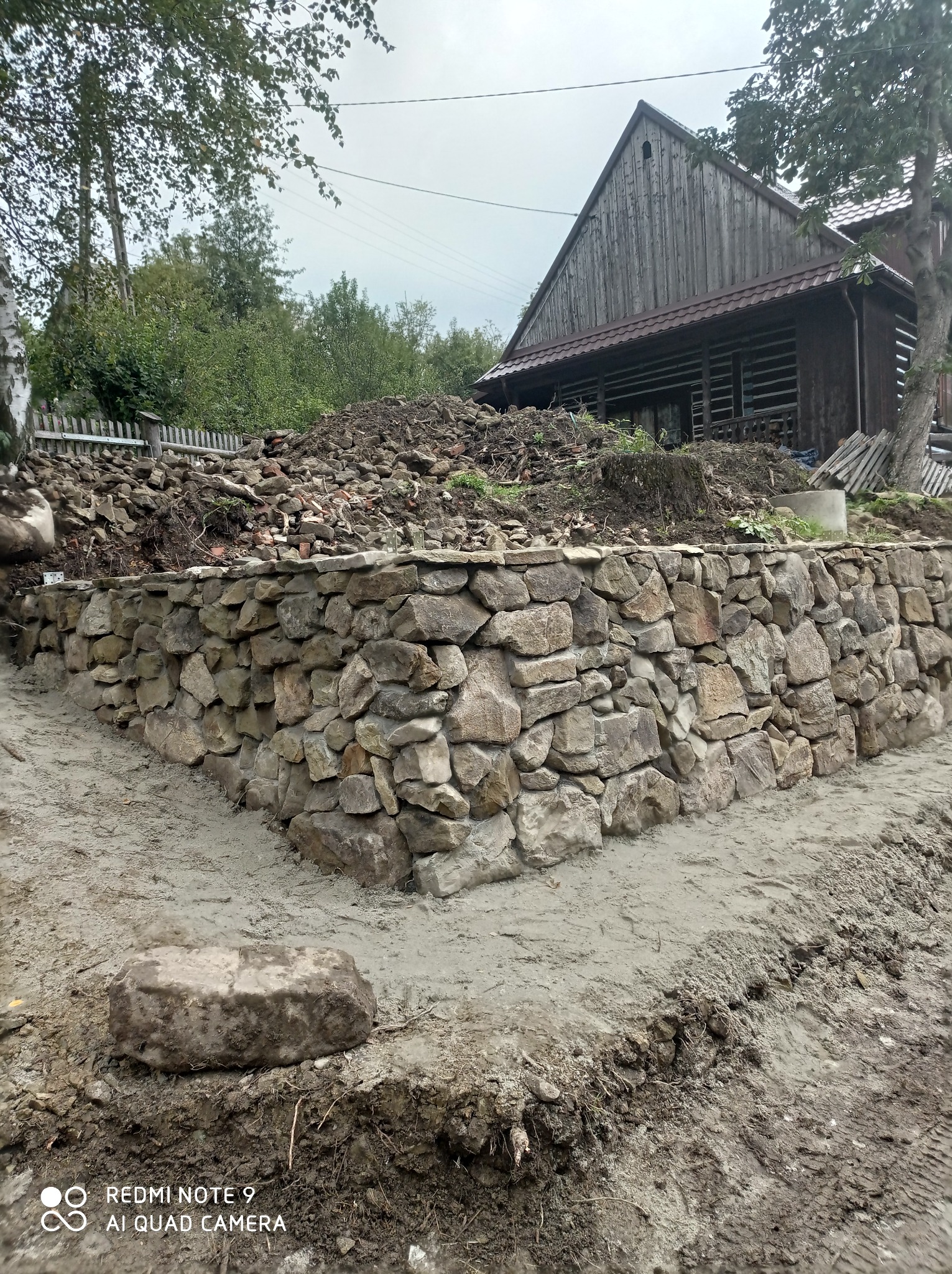 Murek oporowy z kamienia polnego, zabezpieczający skarpę przed domem z drewnianą elewacją, ziemia częściowo usunięta, widoczne ślady prac ziemnych.