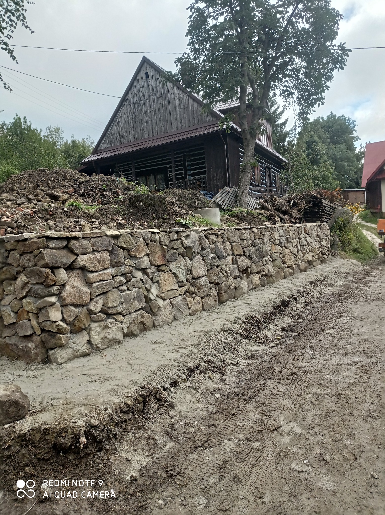 Nowo wybudowany murek oporowy z kamienia polnego przed drewnianym domem z bali, ziemia rozjeżdżona po budowie, widok na wzgórze.