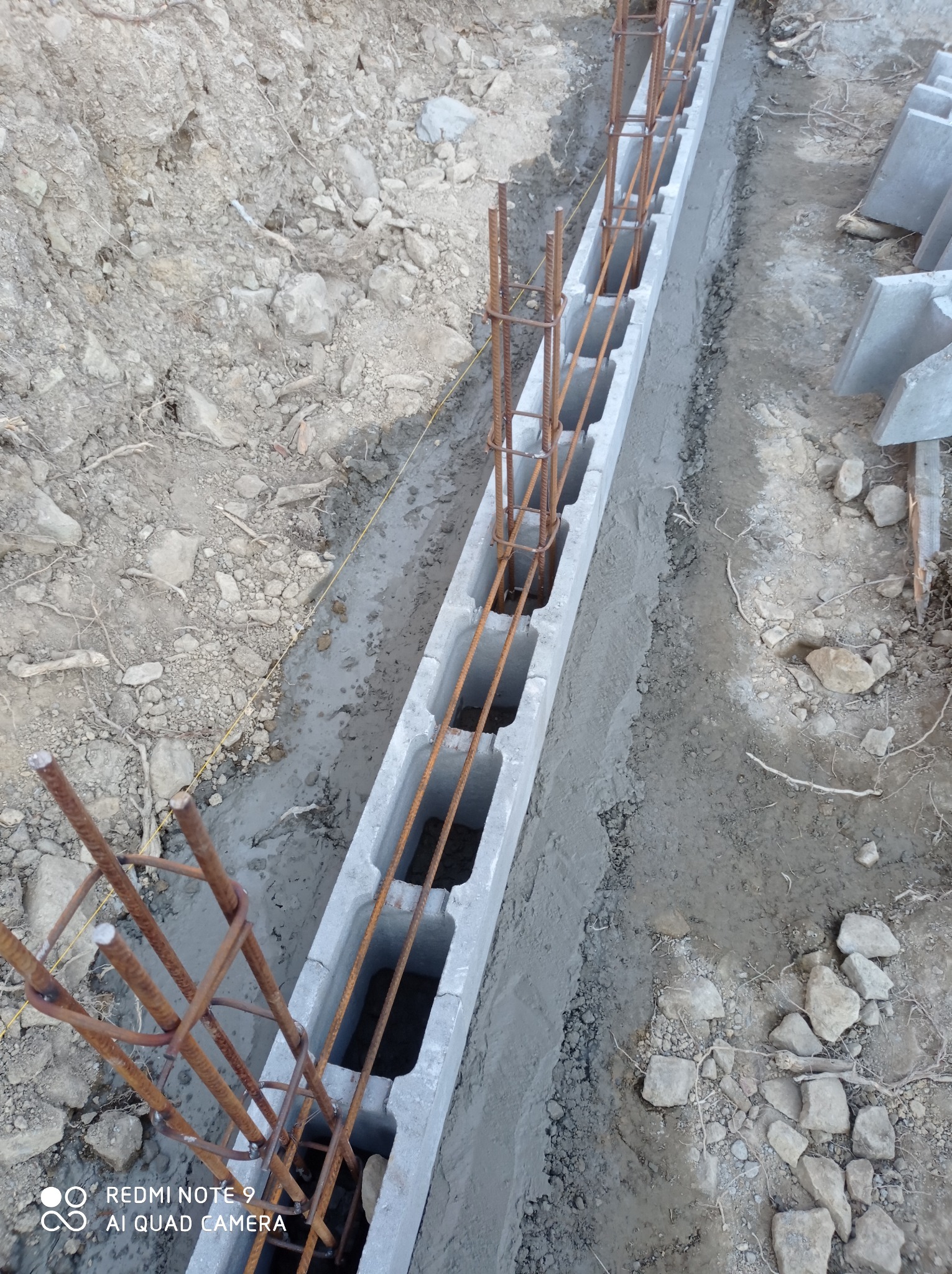 Wykop z betonowymi bloczkami szalunkowymi i zbrojeniem stalowym, widoczne ułożone bloczki obok wykopu, ziemia wokół wykopu.