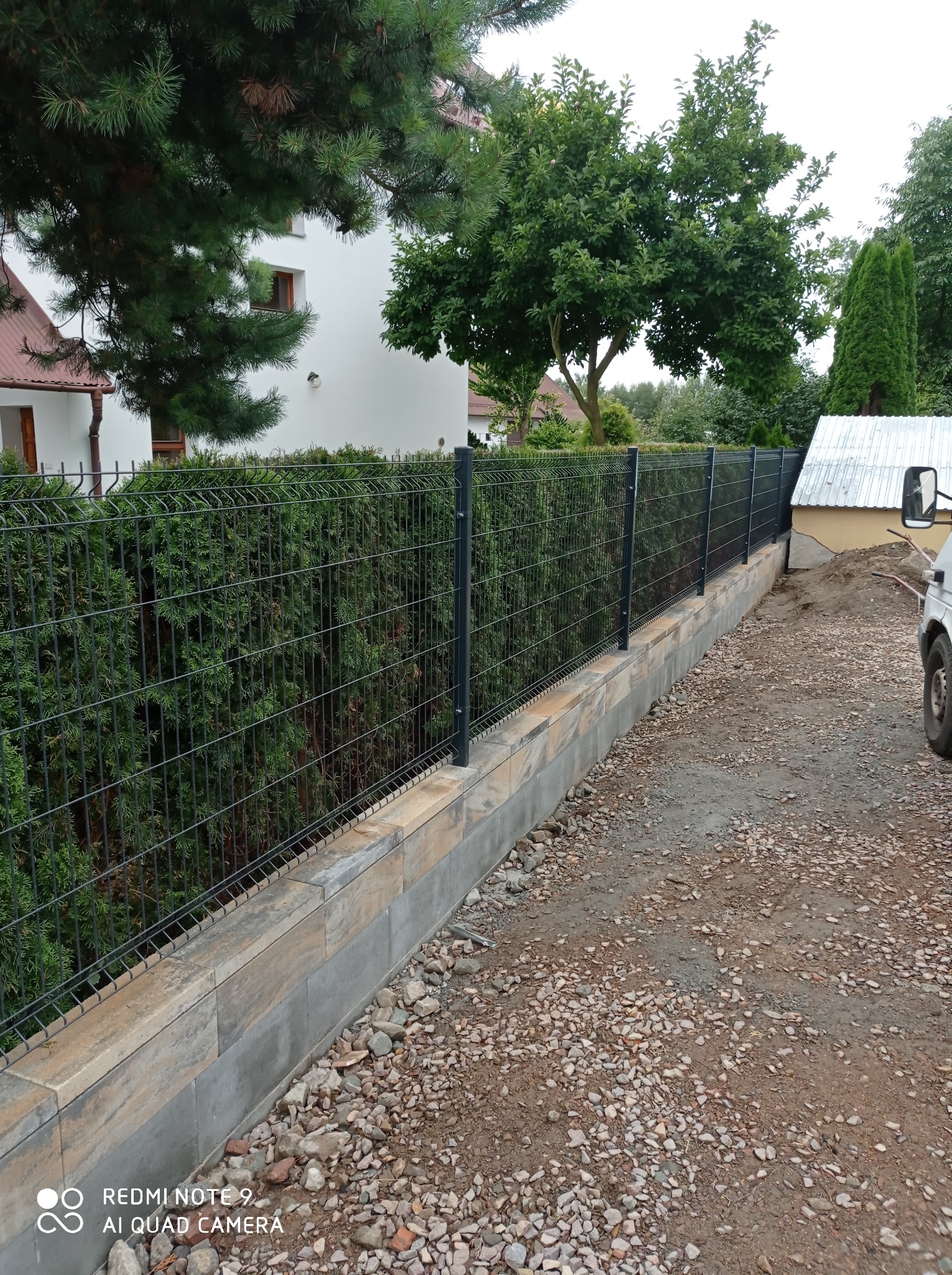 Nowoczesne, grafitowe ogrodzenie panelowe na betonowej podmurówce z widocznymi śladami prac ziemnych, w tle budynek mieszkalny i zieleń.