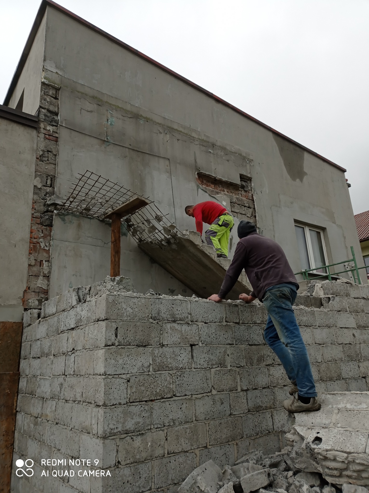 Dwóch pracowników na placu budowy podczas rozbiórki ściany budynku, widoczne fragmenty cegieł, betonowych bloków i zbrojenia, pochmurne niebo w tle.