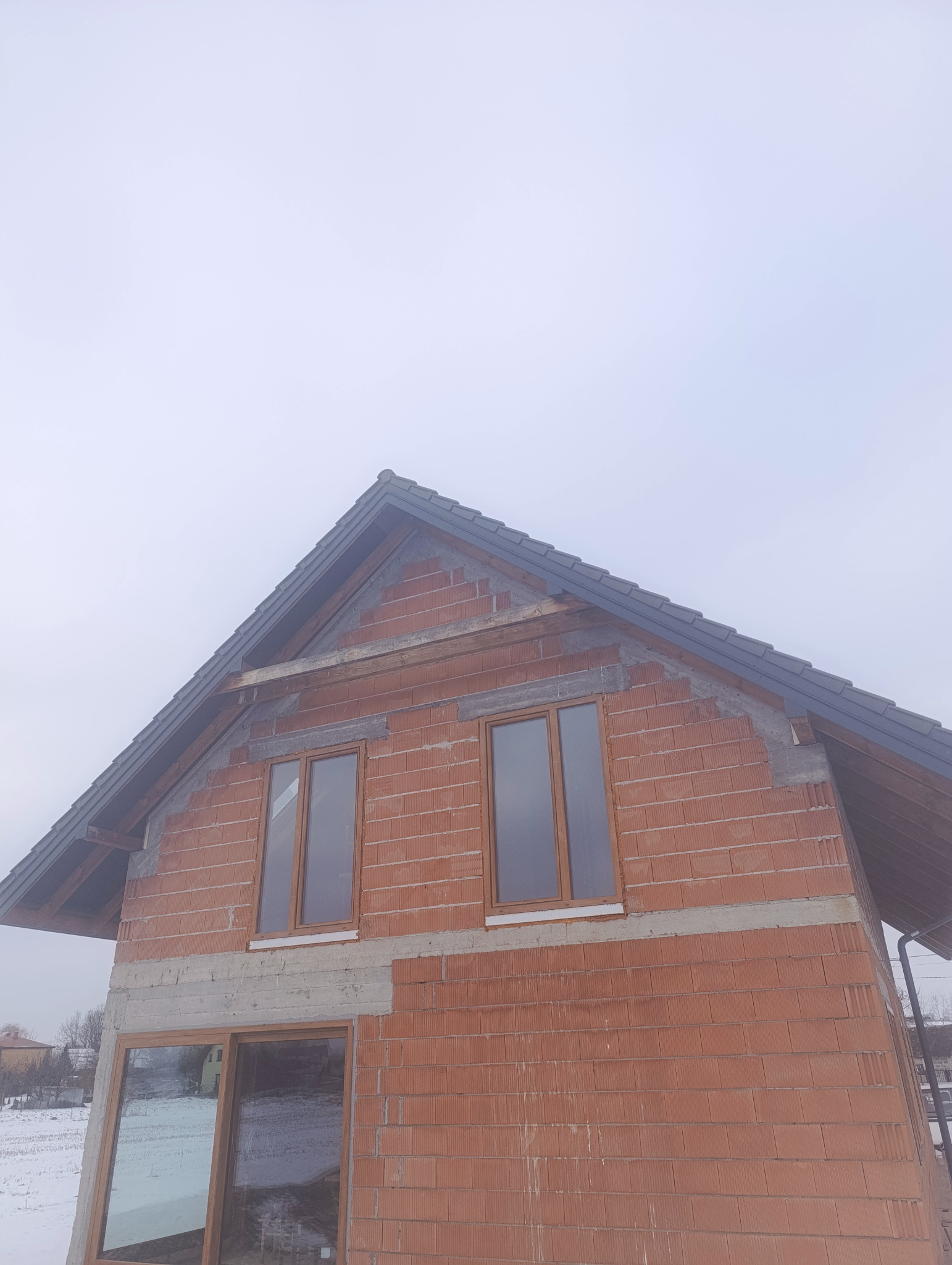 Surowy stan domu z czerwonej cegły z zamontowanymi oknami i szarym dachem, widoczne elementy konstrukcyjne, pochmurne niebo w tle. Budowa domu w trakcie realizacji.