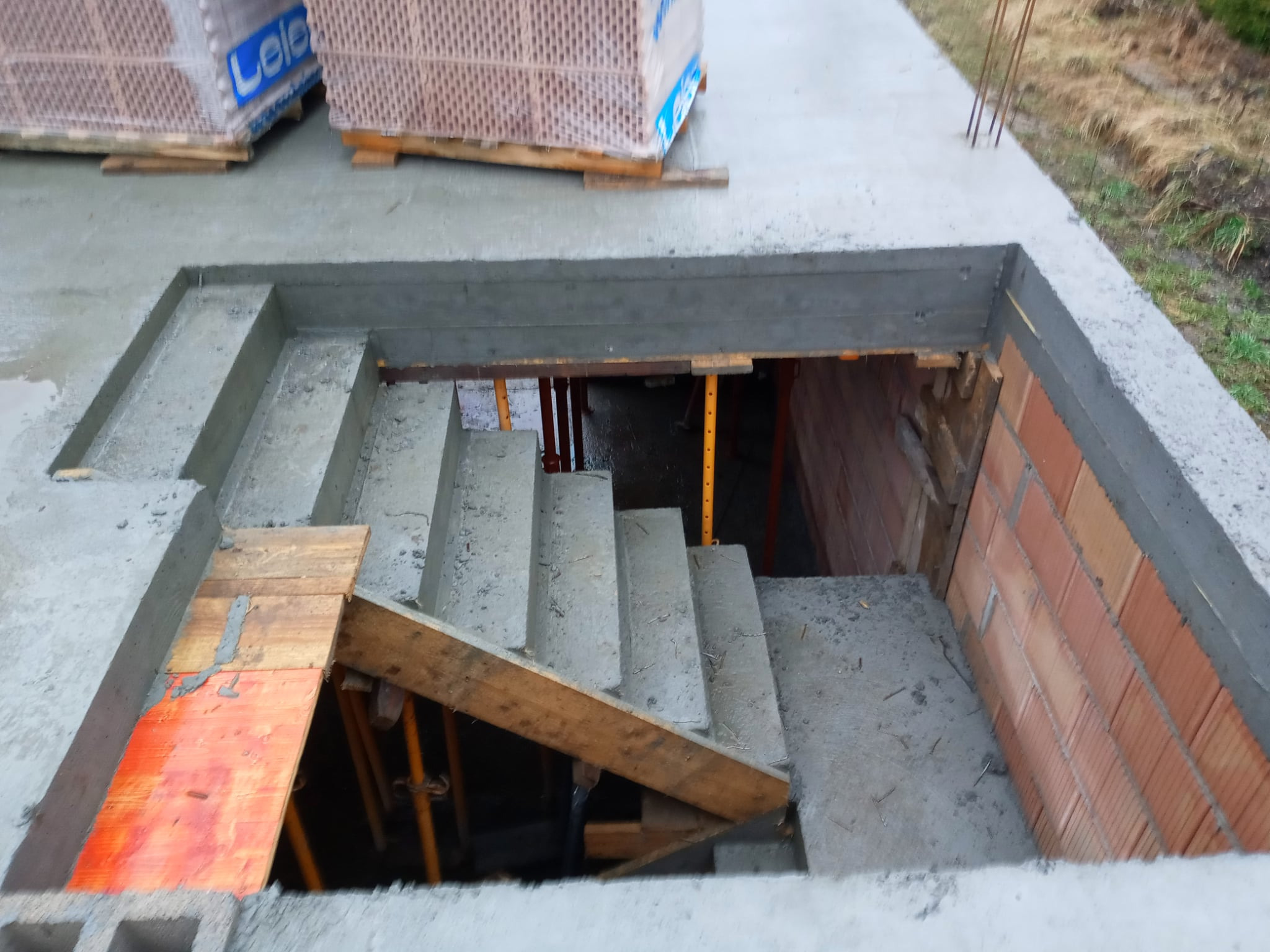 Surowe betonowe schody w trakcie budowy, z widocznym szalunkiem i materiałami budowlanymi. Ujęcie z góry, podkreślające etap prac fundamentowych.