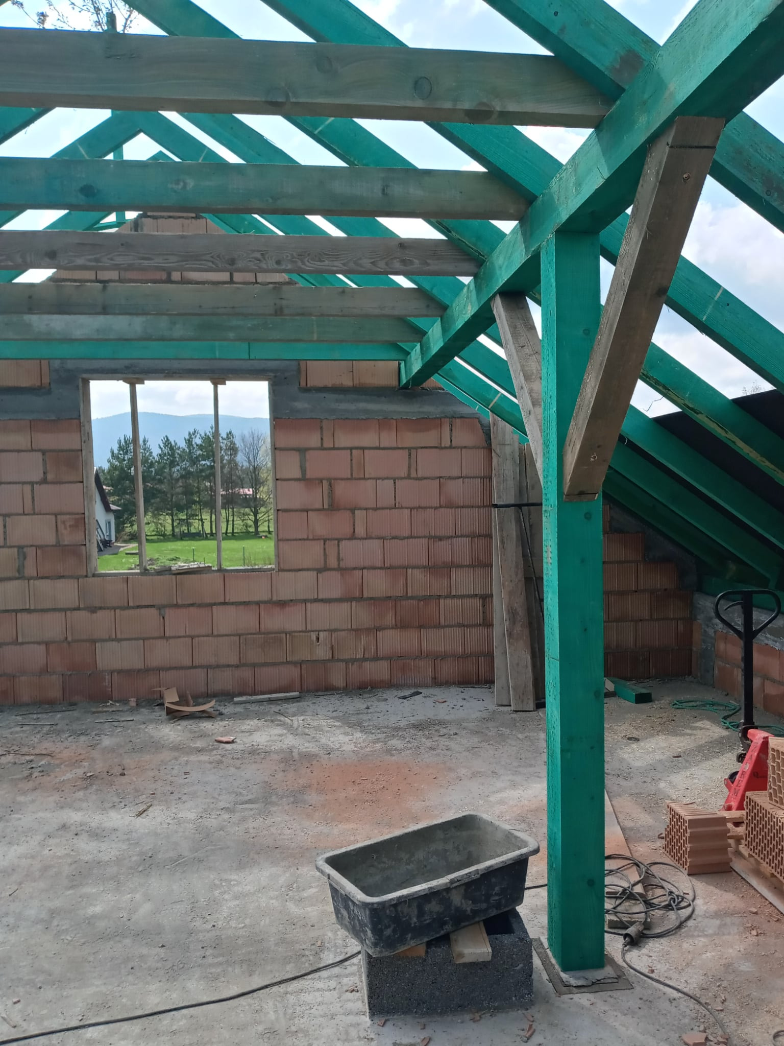 Wnętrze w trakcie murowania ścian z pustaków ceramicznych. Widoczne zielone elementy konstrukcji dachu i krajobraz za oknem. Na podłodze leży kuweta murarska.