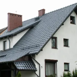 Dom jednorodzinny w Malborku z dachem pokrytym dachówką ceramiczną w odcieniach szarości, widoczny komin z cegły, rynny i okna z brązowymi ramami.