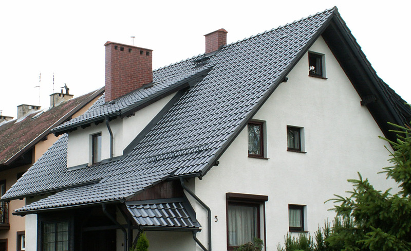 Dom jednorodzinny w Malborku z dachem pokrytym dachówką ceramiczną w odcieniach szarości, widoczny komin z cegły, rynny i okna z brązowymi ramami.