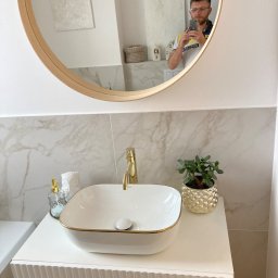 D.B.INVEST - Umywalka nablatowa w białej szafce z frezowanymi frontami i złotymi detalami, złota bateria, marmurowe płytki na ścianie, okrągłe lustro z odbiciem osoby robiącej zdjęcie.