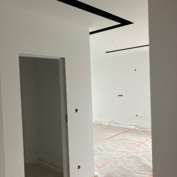 D.B.INVEST - Wnętrze po malowaniu na biało z czarnymi, geometrycznymi elementami dekoracyjnymi na suficie, zabezpieczona folią podłoga i widoczne puszki elektryczne.