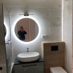 D.B.INVEST - Wykończona łazienka z owalną umywalką nablatową, podświetlanym okrągłym lustrem, zabudowaną toaletą i kabiną prysznicową ze szklaną ścianką, widoczne płytki imitujące beton.