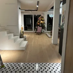 Jasne panele podłogowe w salonie z białymi schodami i choinką w tle, widoczne zza drzwi, z fragmentem drabiny malarskiej w lewym dolnym rogu.