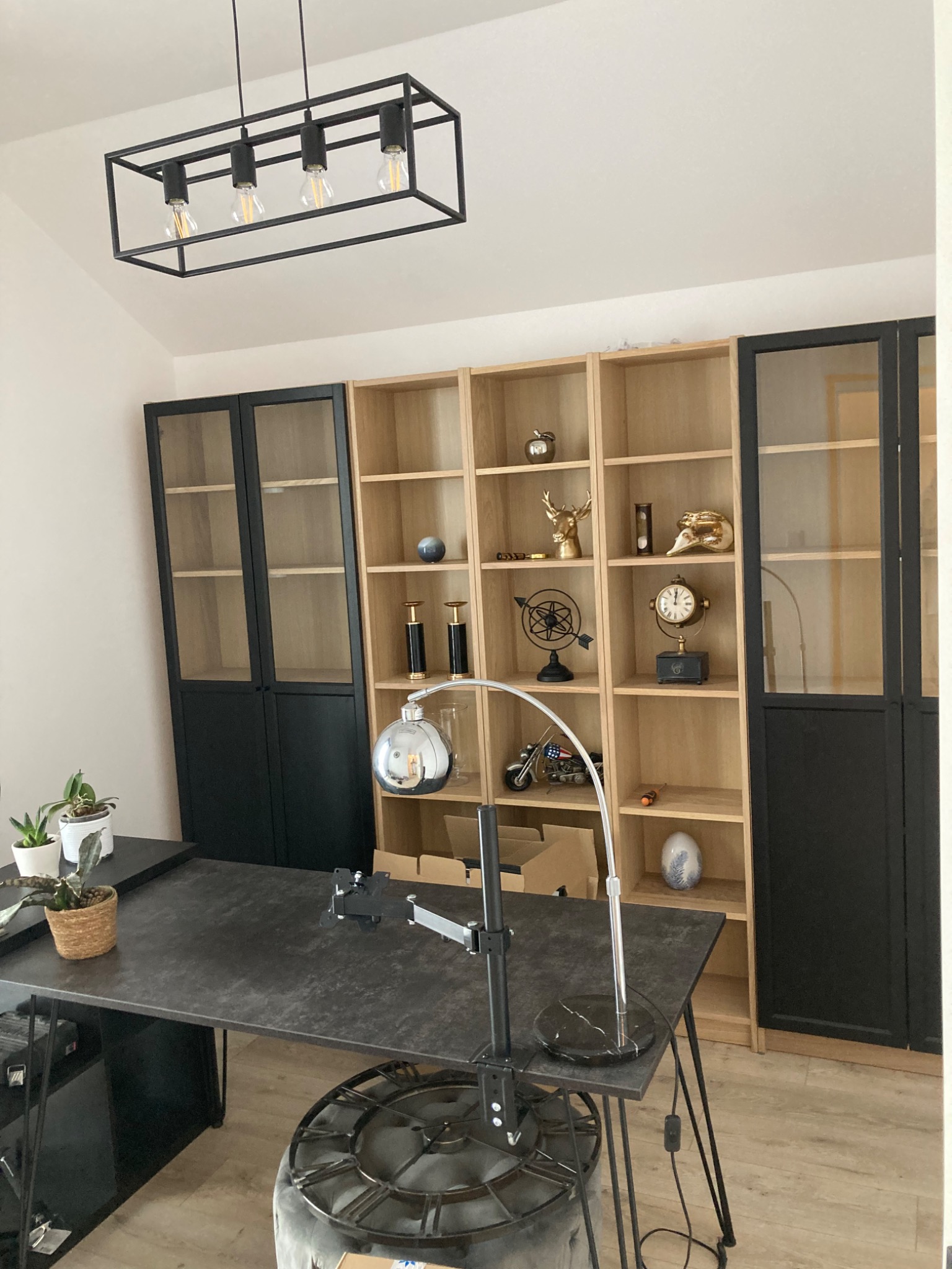 Nowoczesne biuro z czarnym blatem, regałami z jasnego drewna i czarnymi szafkami z przeszklonymi drzwiami, oświetlone minimalistyczną lampą wiszącą i lampką biurkową.