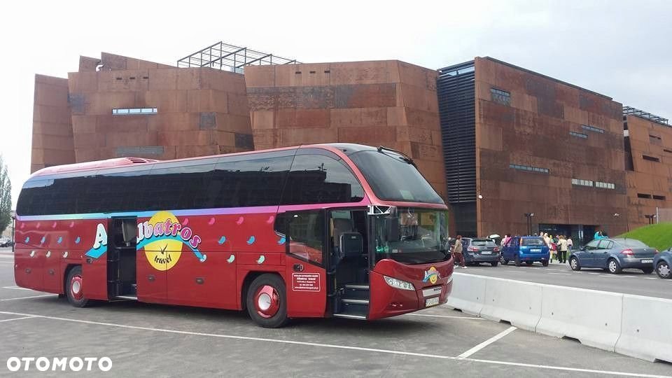 Czerwony autokar turystyczny z otwartymi drzwiami i kolorowym logo 'Albatros Travel' na tle nowoczesnego budynku z rdzawego metalu.