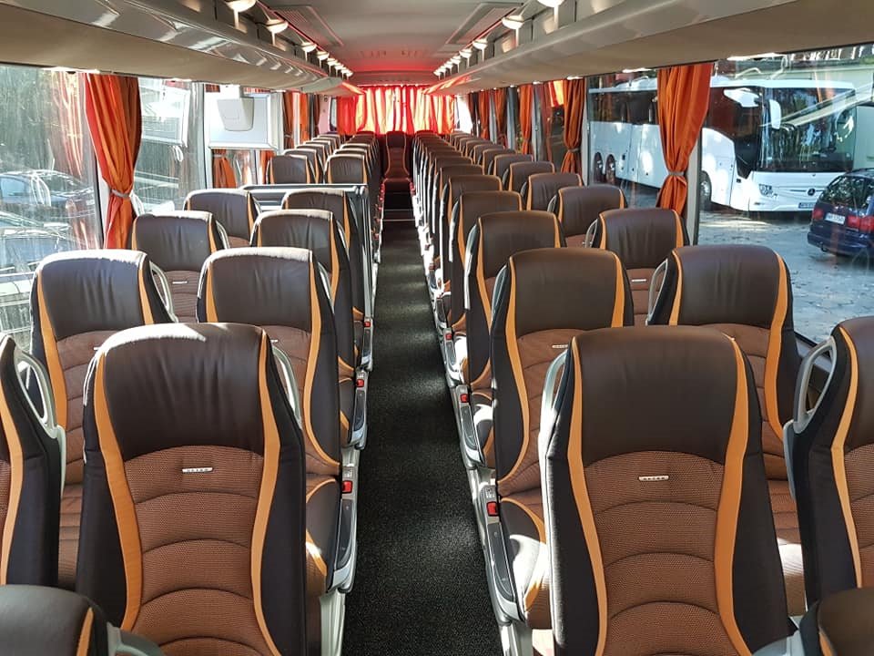 Wnętrze autobusu turystycznego z brązowo-pomarańczowymi fotelami i zasłonami, widok na puste siedzenia wzdłuż korytarza, na zewnątrz widoczne inne autobusy.