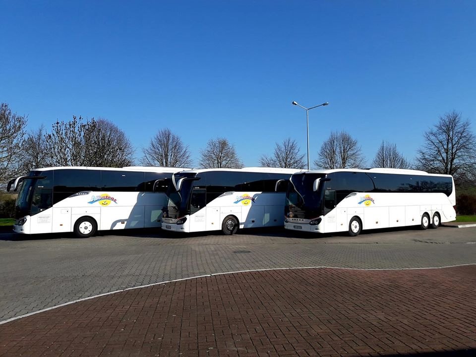 Trzy białe autokary turystyczne z logo firmy na boku, stojące na parkingu w słoneczny dzień.