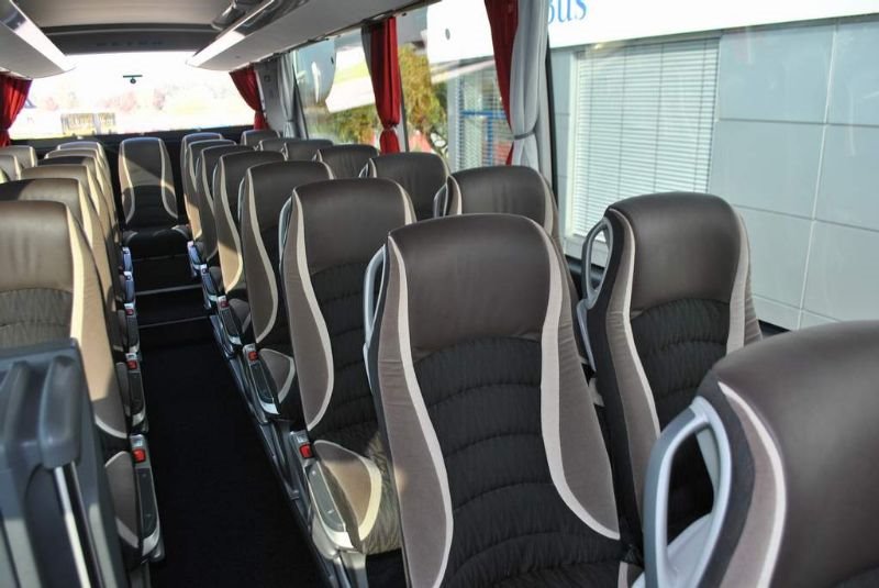 Wnętrze autobusu turystycznego z rzędami foteli obitych ciemną tapicerką z szarymi wstawkami, widok z perspektywy pasażera, zasłony w oknach.