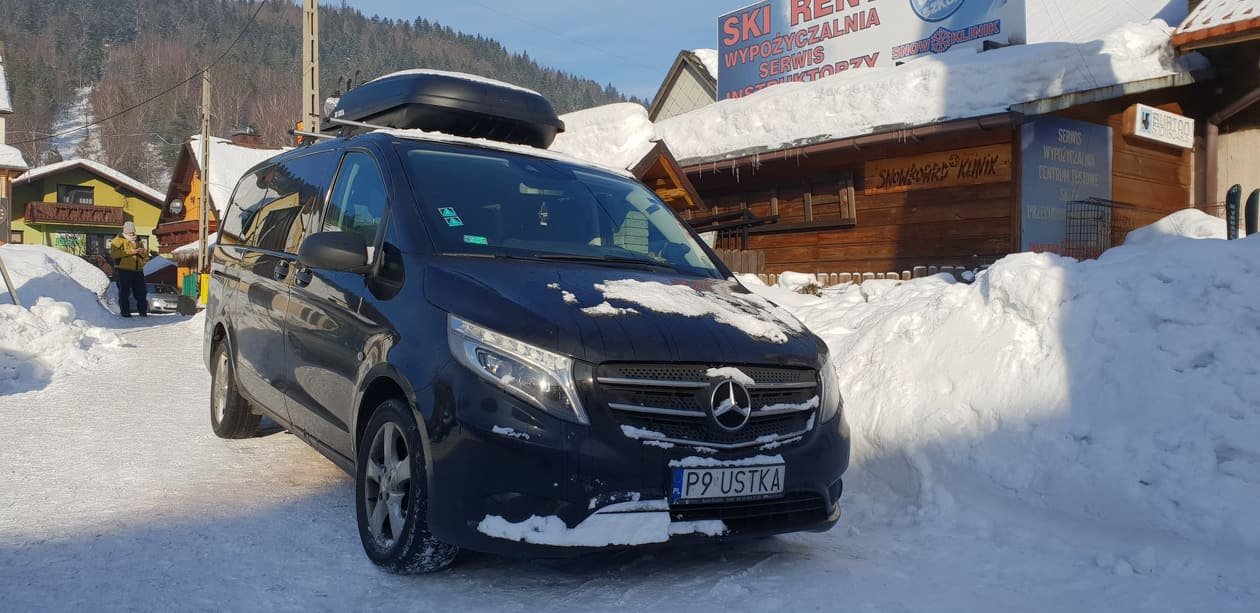 Czarny Mercedes V-klasa z bagażnikiem dachowym na tle zimowego krajobrazu i budynku wypożyczalni sprzętu narciarskiego.