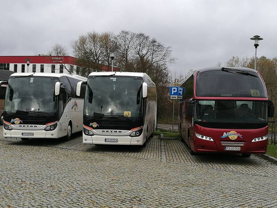 Trzy autokary (dwa białe marki Setra i jeden czerwony) zaparkowane na brukowanym placu, widok z przodu, pochmurny dzień.