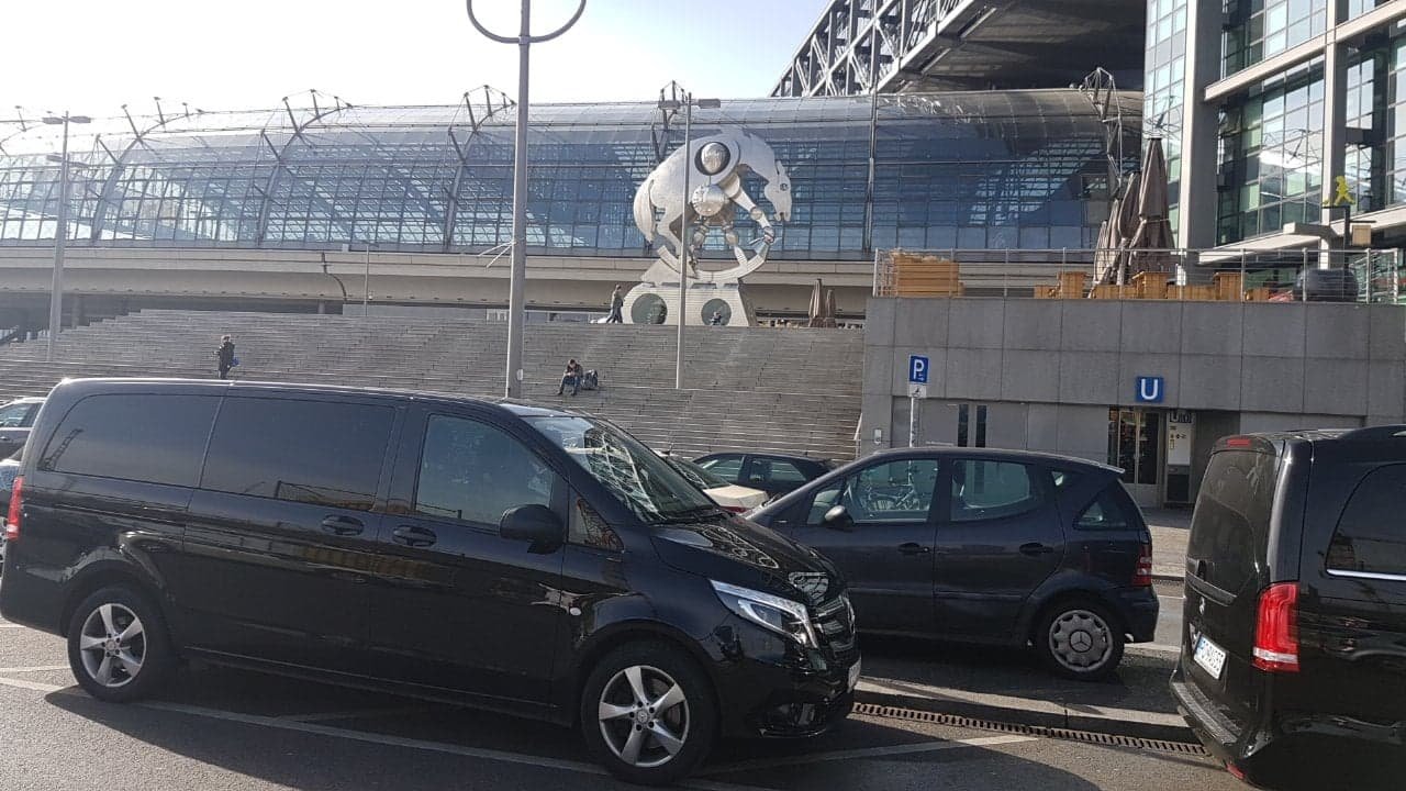 Czarne busy na parkingu przed budynkiem z przeszklonym dachem i rzeźbą konia, w tle wejście do metra.