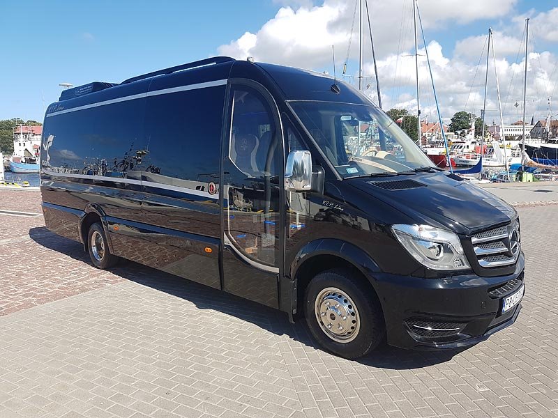 Czarny, luksusowy Mercedes Sprinter VIP Line na tle portu z jachtami, odbicia w karoserii, kostka brukowa.