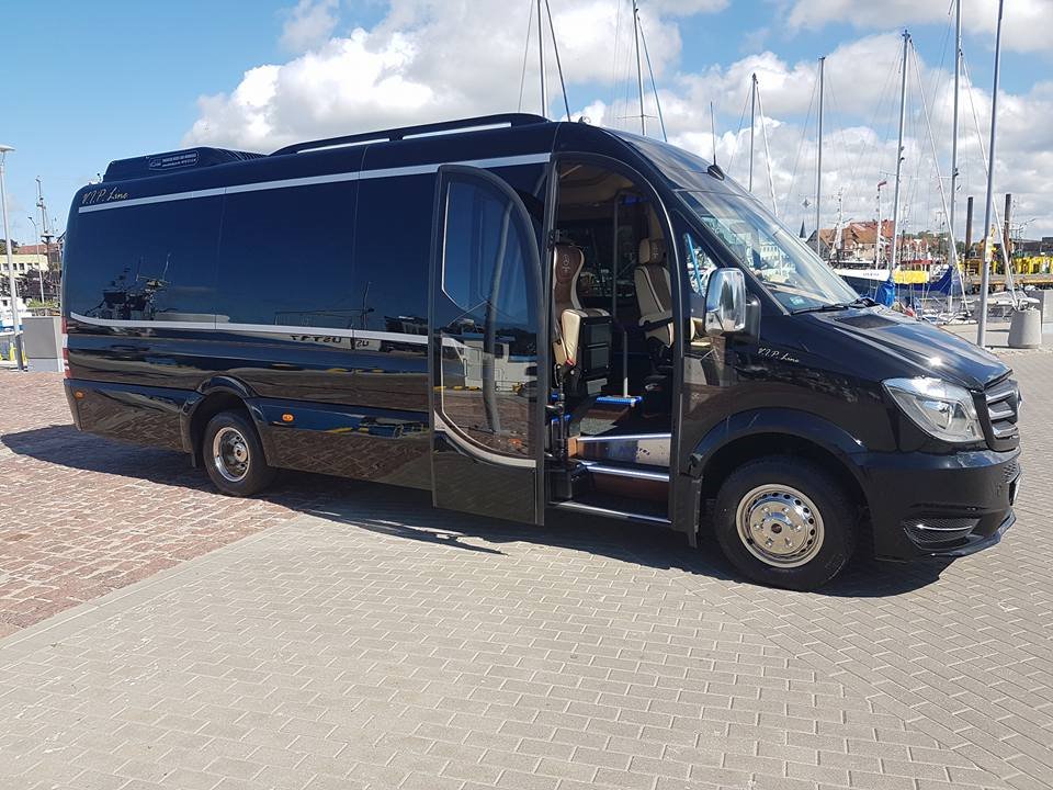 Czarny, luksusowy bus z otwartymi drzwiami na tle portu jachtowego z masztami i błękitnym niebem.