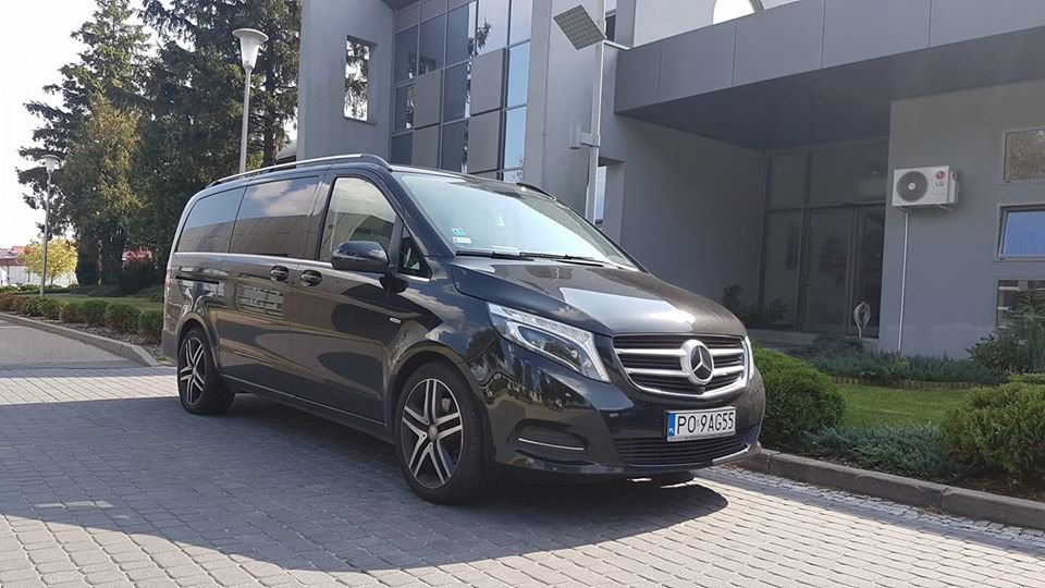 Czarny Mercedes-Benz V-klasy zaparkowany przed nowoczesnym budynkiem biurowym z szarymi panelami i dużymi oknami, widoczna tablica rejestracyjna PO 9AG55.