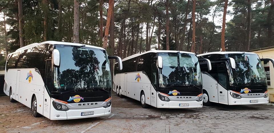 Trzy białe autokary marki Setra zaparkowane na placu, widoczne tablice rejestracyjne i logo firmy z kolorowym rysunkiem słońca.