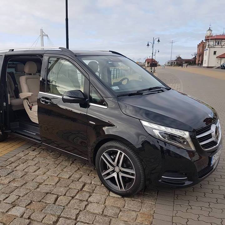 Czarny van Mercedes V-klasy z otwartymi drzwiami przesuwnymi, beżowe skórzane wnętrze, na brukowanym chodniku, w tle nadmorski krajobraz z latarnią morską i mostem.