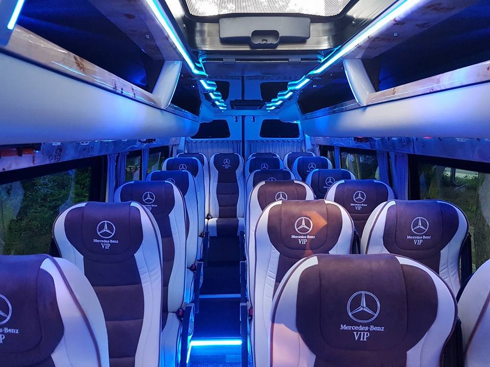 Wnętrze luksusowego busa Mercedes-Benz VIP z podświetleniem LED, widok na rzędy foteli z logo firmy.