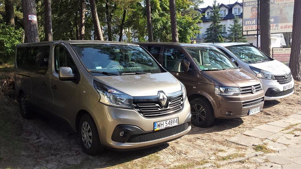 Trzy busy: beżowy Renault Trafic z tablicą WH 54894, brązowy Volkswagen Transporter z tablicą P2 USTRA i biały bus, zaparkowane obok siebie na parkingu z drzewami w tle.