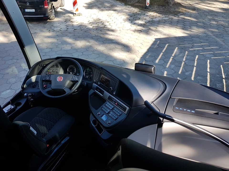 Wnętrze kabiny kierowcy autobusu Setra, widok na deskę rozdzielczą, kierownicę z logo i fotel kierowcy, na zewnątrz widoczny samochód i chodnik.