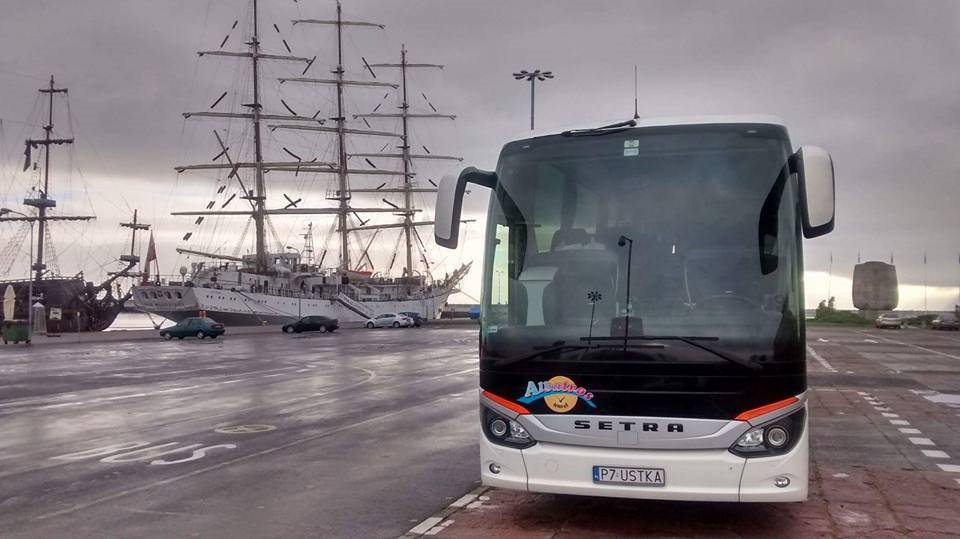 Biały autokar marki Setra z logo firmy na tle dwóch żaglowców w porcie przy pochmurnym niebie.