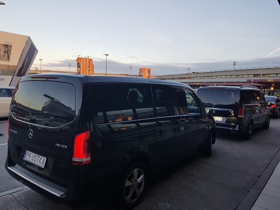Ciemny minibus Mercedes 116 CDI z polską rejestracją P9 USTKA, stojący w kolejce na lotnisku, widok z tyłu, w tle budynek lotniska i inne busy.