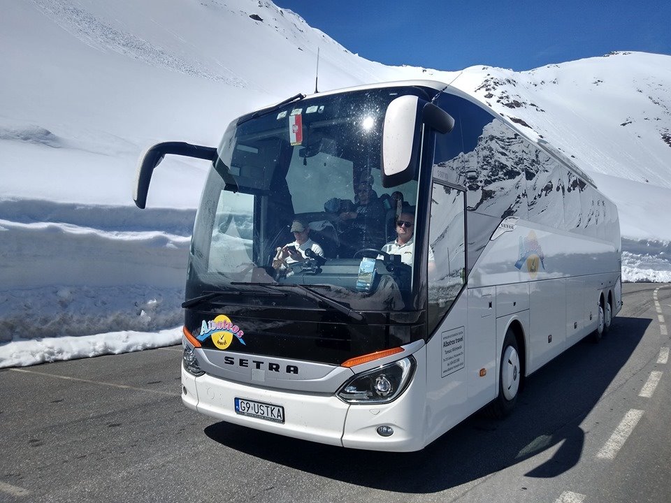 Biały autokar SETRA z logiem Albatros Travel na tle ośnieżonych gór, kierowca w okularach i pasażerowie wewnątrz pojazdu, tablica rejestracyjna G9 USTKA.