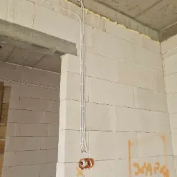 Białe kable elektryczne przymocowane do ściany z betonu komórkowego, pomarańczowa puszka elektryczna widoczna u dołu, ściana częściowo pokryta pianką montażową.