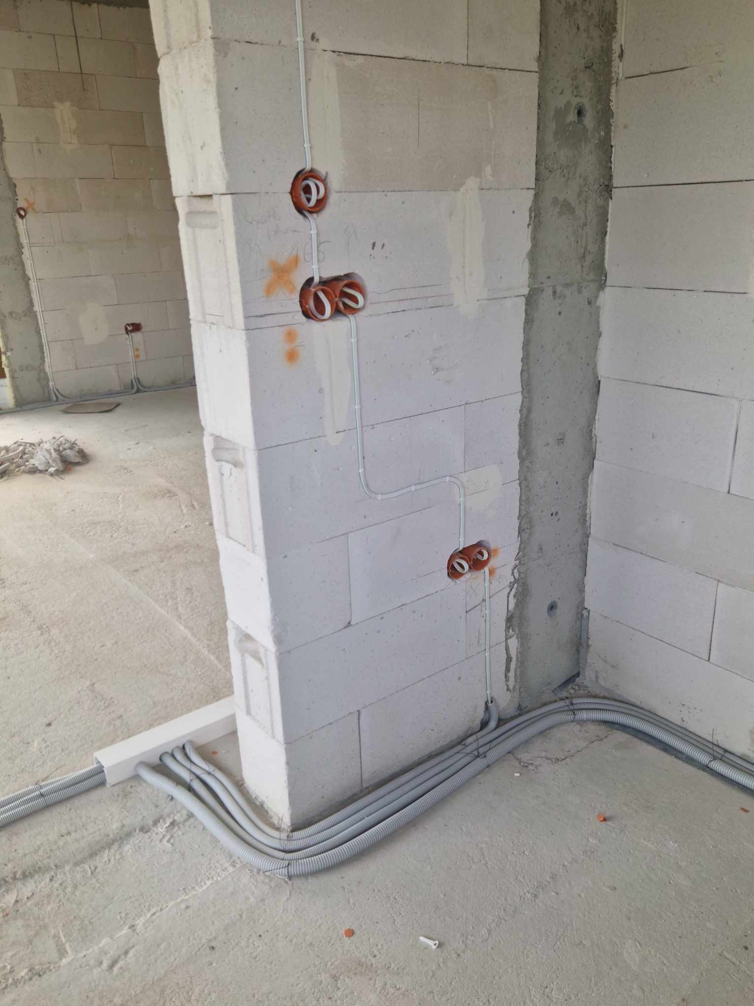 Wnętrze w trakcie budowy z widocznymi przewodami elektrycznymi w ścianach z betonu komórkowego i szarymi rurkami karbowanymi prowadzonymi wzdłuż podłogi, zabezpieczonymi białym korytkiem.